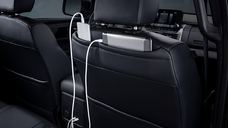 Innenansicht des Mitsubishi L200 von hinten: Blick auf die Rückseiten der Vordersitze mit integrierter Halterung, in der ein mobiles Gerät steckt und per Ladekabel angeschlossen ist – zeigt die praktischen Ablage- und Lademöglichkeiten für Passagiere im Fond.