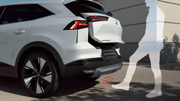 Rückansicht eines weißen Grandis Select SUV mit geöffneter Heckklappe; eine stilisierte Person aktiviert per Fußbewegung den Sensor unter dem Heckstoßfänger für das berührungslose Öffnen des Kofferraums.