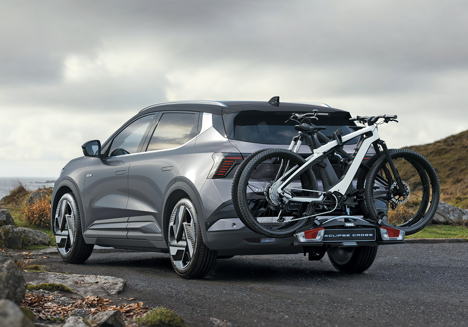 Graue Eclipse Cross Intro Edition in schräger Heckansicht, geparkt auf einer Küstenstraße mit Felsen und Meer im Hintergrund; am Heck ist ein Fahrradträger mit zwei modernen Fahrrädern montiert – symbolisiert den aktiven Lifestyle und die Eignung für Ausflüge und Outdoor-Abenteuer.