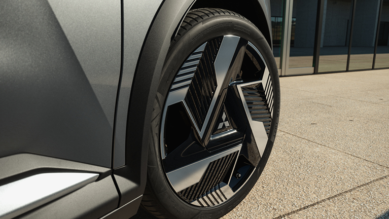 Nahaufnahme des Vorderrads der Eclipse Cross Intro Edition mit auffälligen, aerodynamisch gestalteten zweifarbigen Leichtmetallfelgen vor moderner Architektur – unterstreicht den futuristischen, hochwertigen Look des E-SUV.