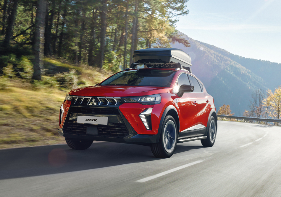 Roter Mitsubishi ASX Select mit Dachbox fährt dynamisch auf einer kurvenreichen Bergstraße, umgeben von Wald und Bergen bei sonnigem Wetter – symbolisiert Fahrspaß und Vielseitigkeit für Ausflüge.