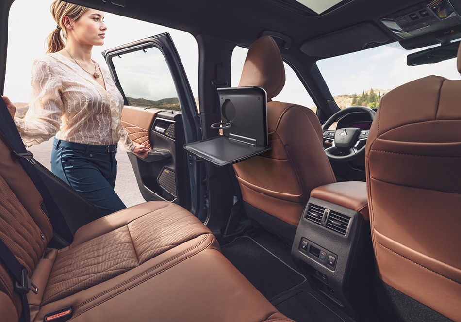 Blick in den braunen Leder-Innenraum eines Mitsubishi Outlander; eine Frau steht an der geöffneten hinteren Tür, vor dem Rücksitz ist an der Rückenlehne des Vordersitzes ein Klapptisch mit Getränkehalter ausgeklappt.