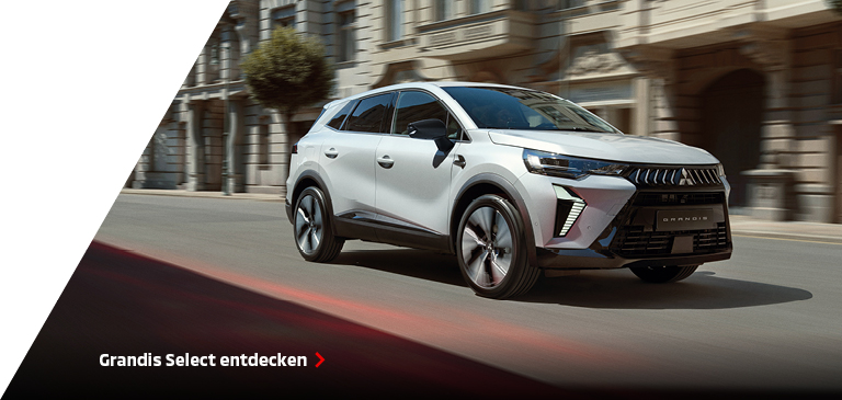 Weißer Mitsubishi Grandis Select fährt durch eine städtische Straße mit klassischen Fassaden im Hintergrund, hervorgehoben als komfortabler Begleiter für den Alltag in der Stadt.