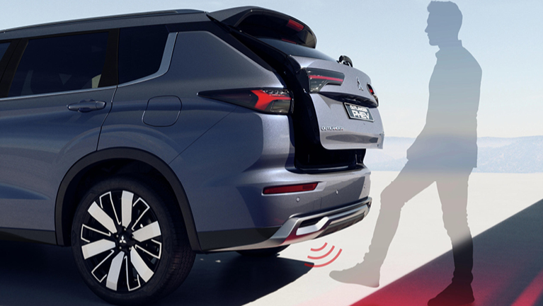 Rückansicht eines grauen Outlander Plug-in Hybrid Select, dessen Heckklappe sich automatisch öffnet, während eine Person seitlich am Fahrzeug mit einer Fußbewegung den berührungslosen Öffnungssensor am Heck betätigt.